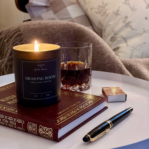 Candle labeled 'Drawing Room' on a book with a glass of whiskey, pen, and lighter on a table.