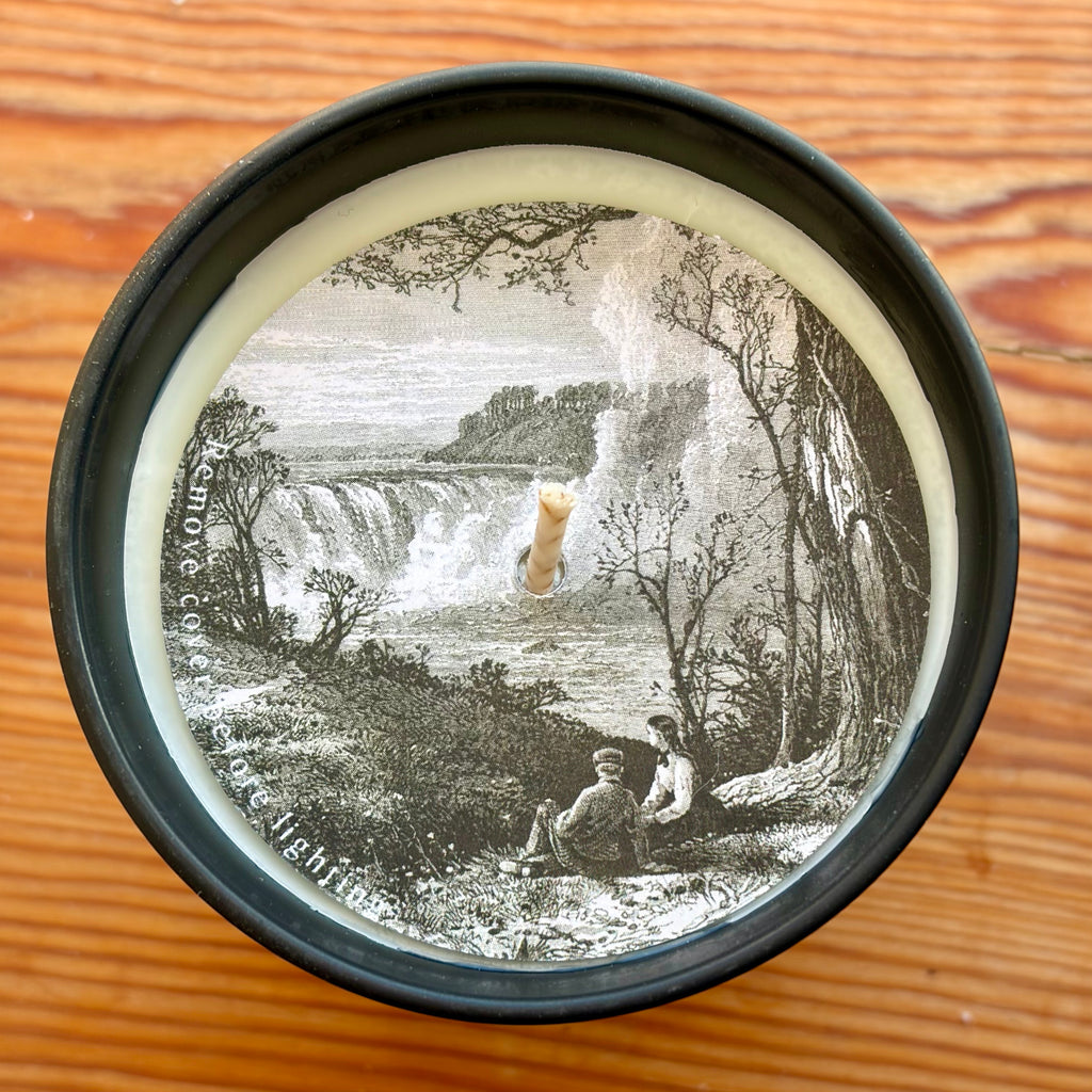 Round container lid with vintage-style illustration of a waterfall and people on a wooden surface