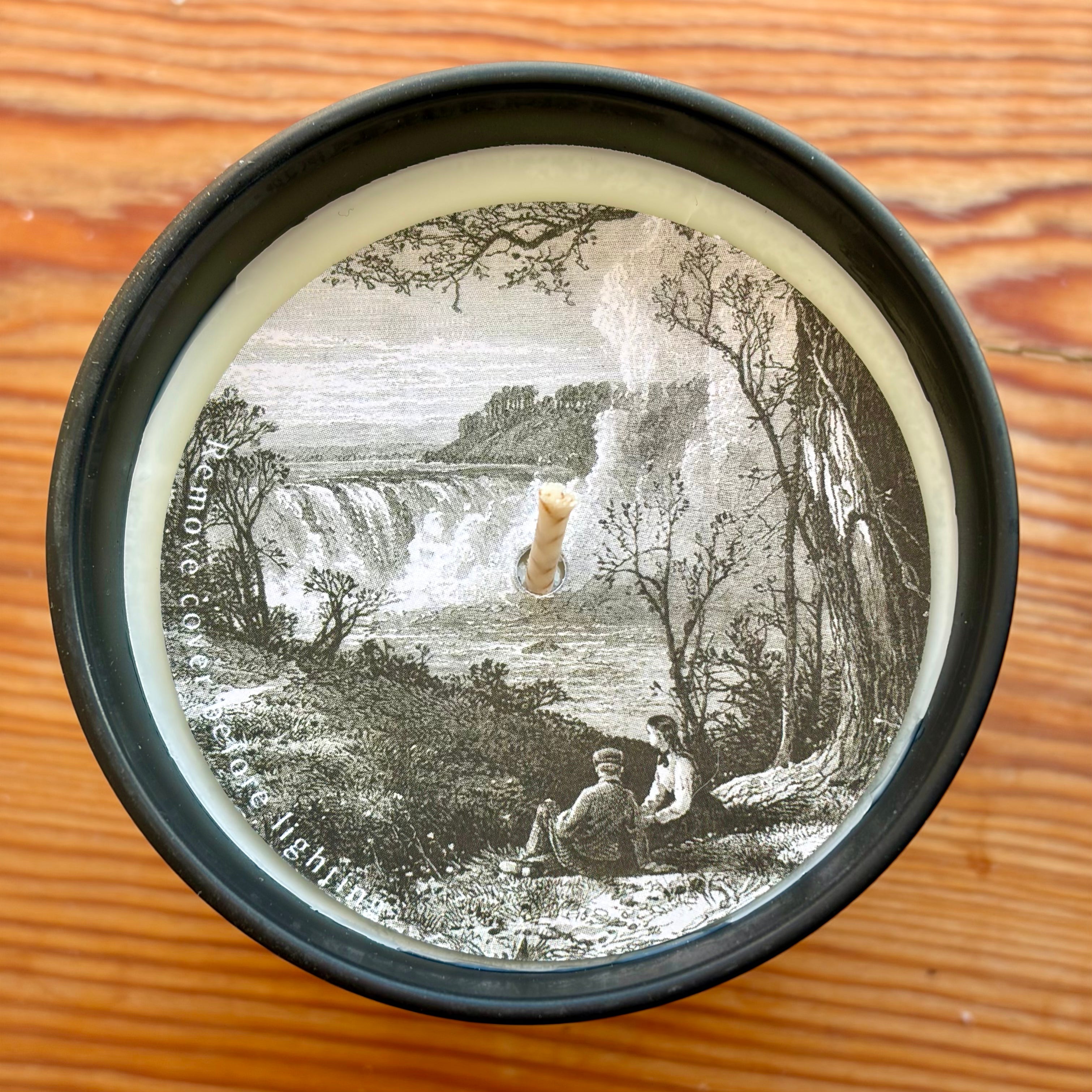 Round container lid with vintage-style illustration of a waterfall and people on a wooden surface